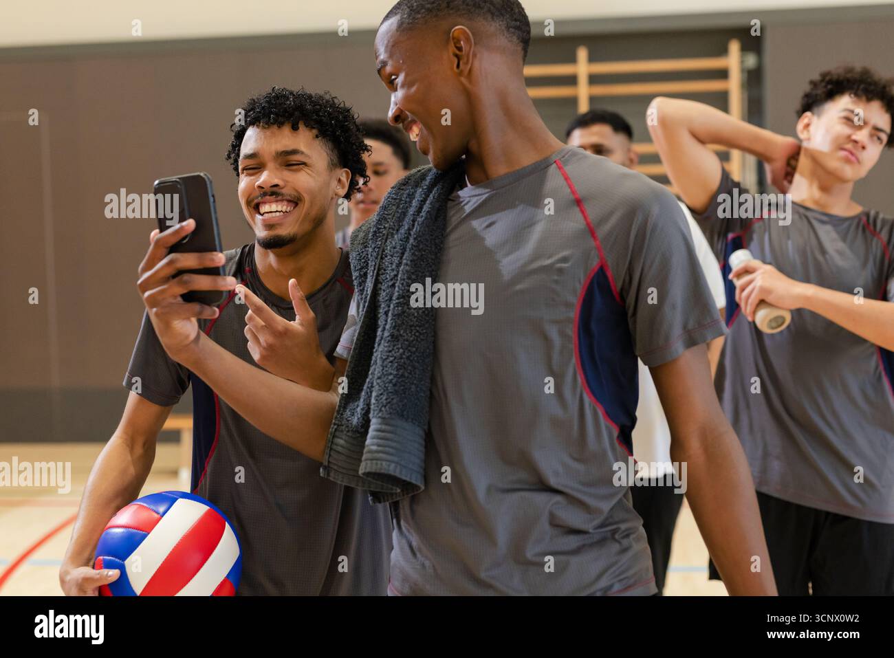 Divers partenaires masculins de volleyball interagissant sur smartphone dans la salle de gym de l'école avec balle et serviette Banque D'Images