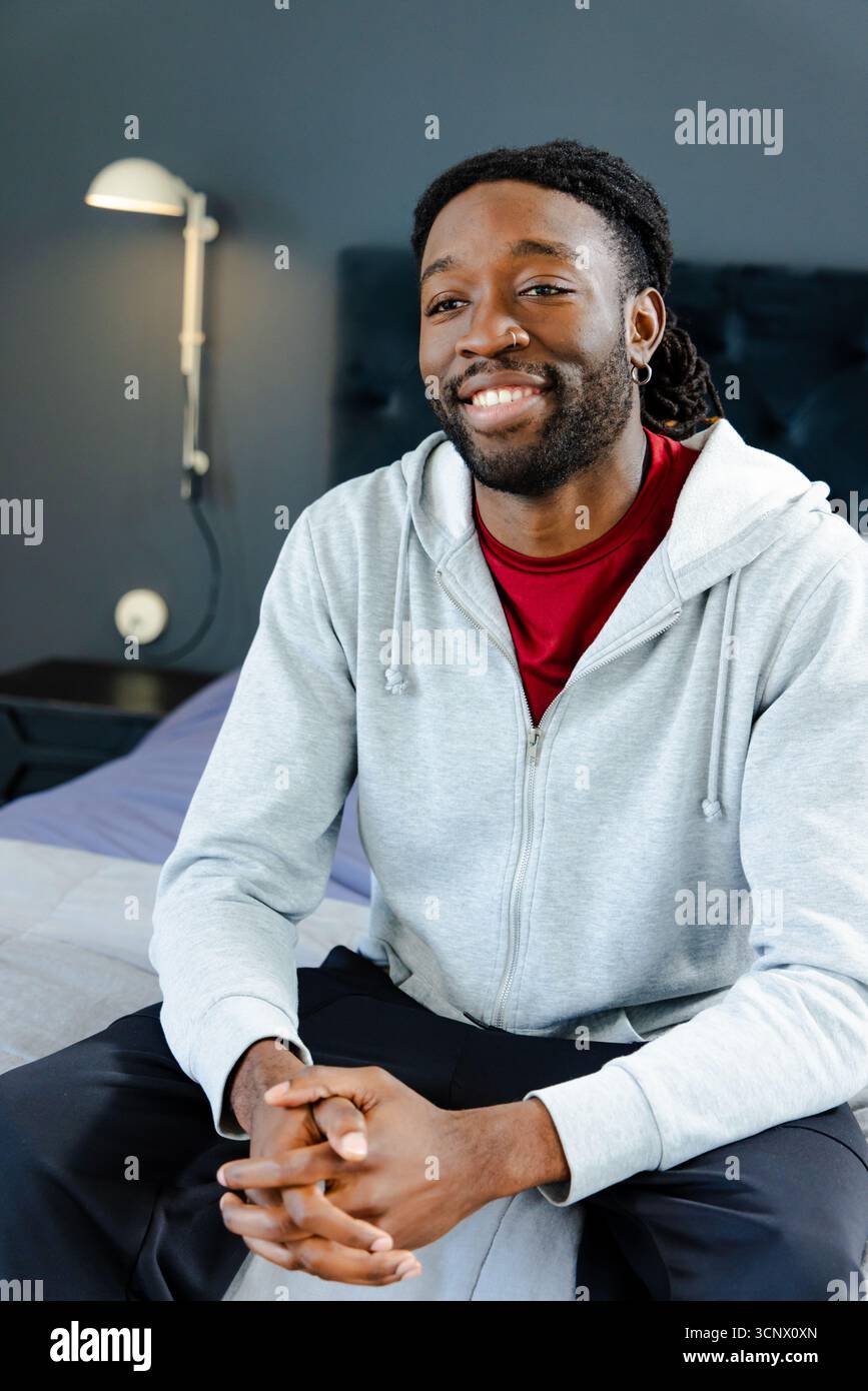 Homme afro-américain assis sur le lit de la chambre avec lampe de tête de lit et table de nuit portant un sweat à capuche gris Banque D'Images