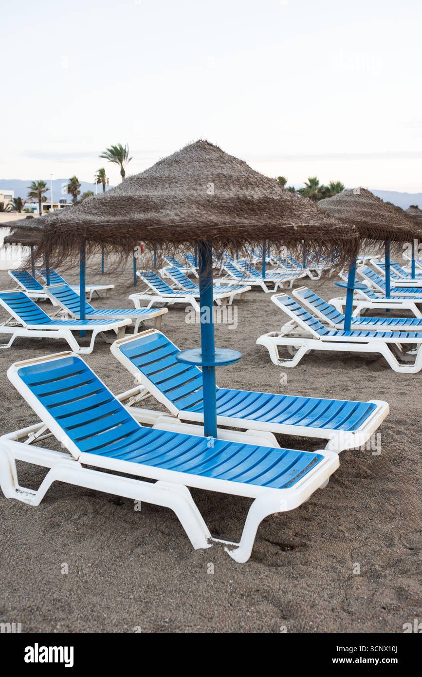 Des chaises longues bleues avec parasols de paille sont installées sur le rivage de sable, invitant à la détente et au plaisir dans un environnement balnéaire tranquille rempli de chaleur Banque D'Images