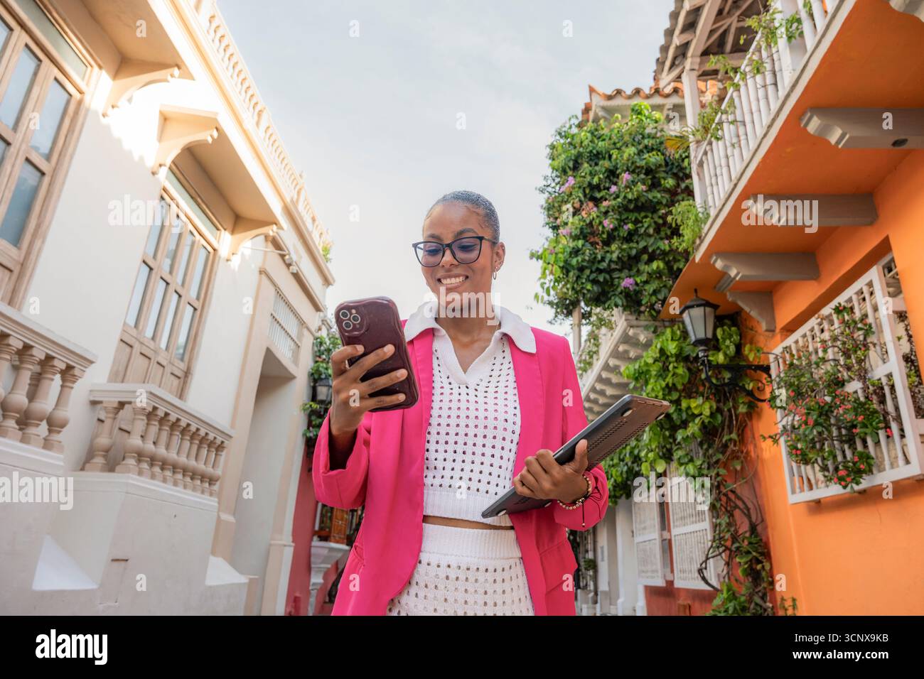Femme d'affaires noire confiante dans un costume rose vif utilise son smartphone et tient une tablette tout en souriant dans une charmante rue de Cartagena de Indias, Banque D'Images