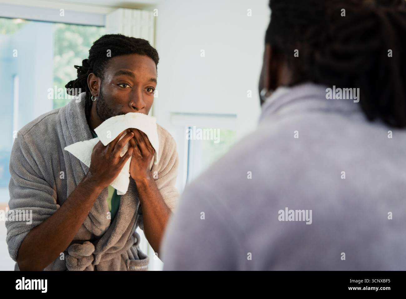 Homme afro-américain face au miroir séchant le visage avec serviette blanche à l'évier de la salle de bain dans un peignoir gris Banque D'Images