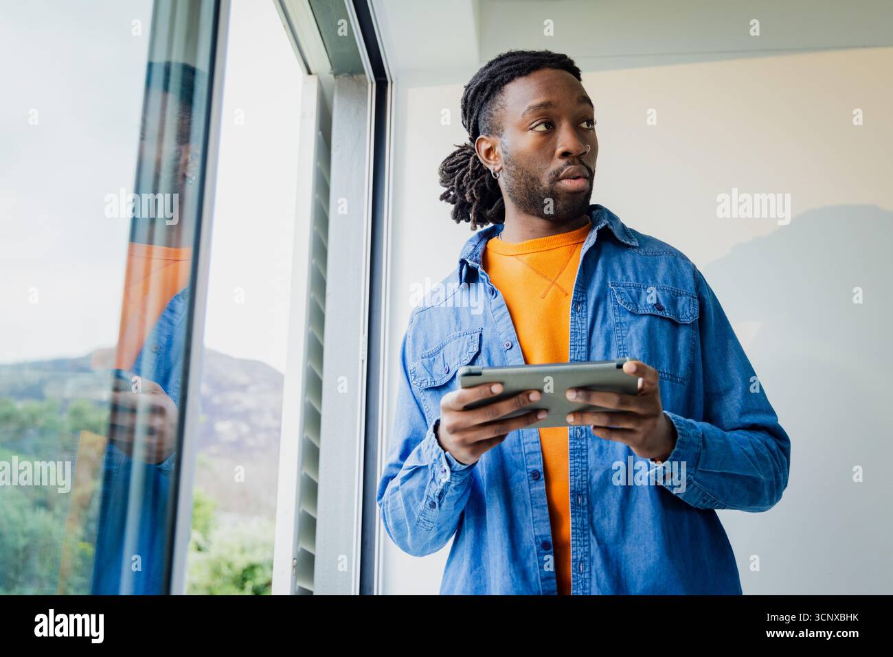 Homme afro-américain debout par fenêtre en verre dans l'intérieur moderne tenant tablette numérique, espace de copie Banque D'Images
