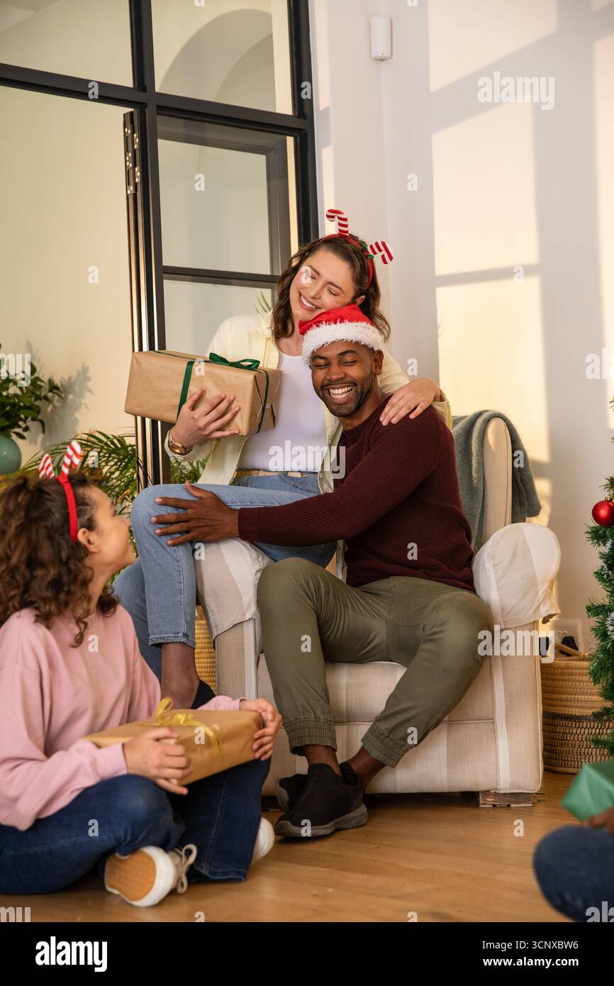 Famille diversifiée échangeant des cadeaux dans le salon confortable portant chapeau de père noël, bandeaux de canne de bonbons Banque D'Images