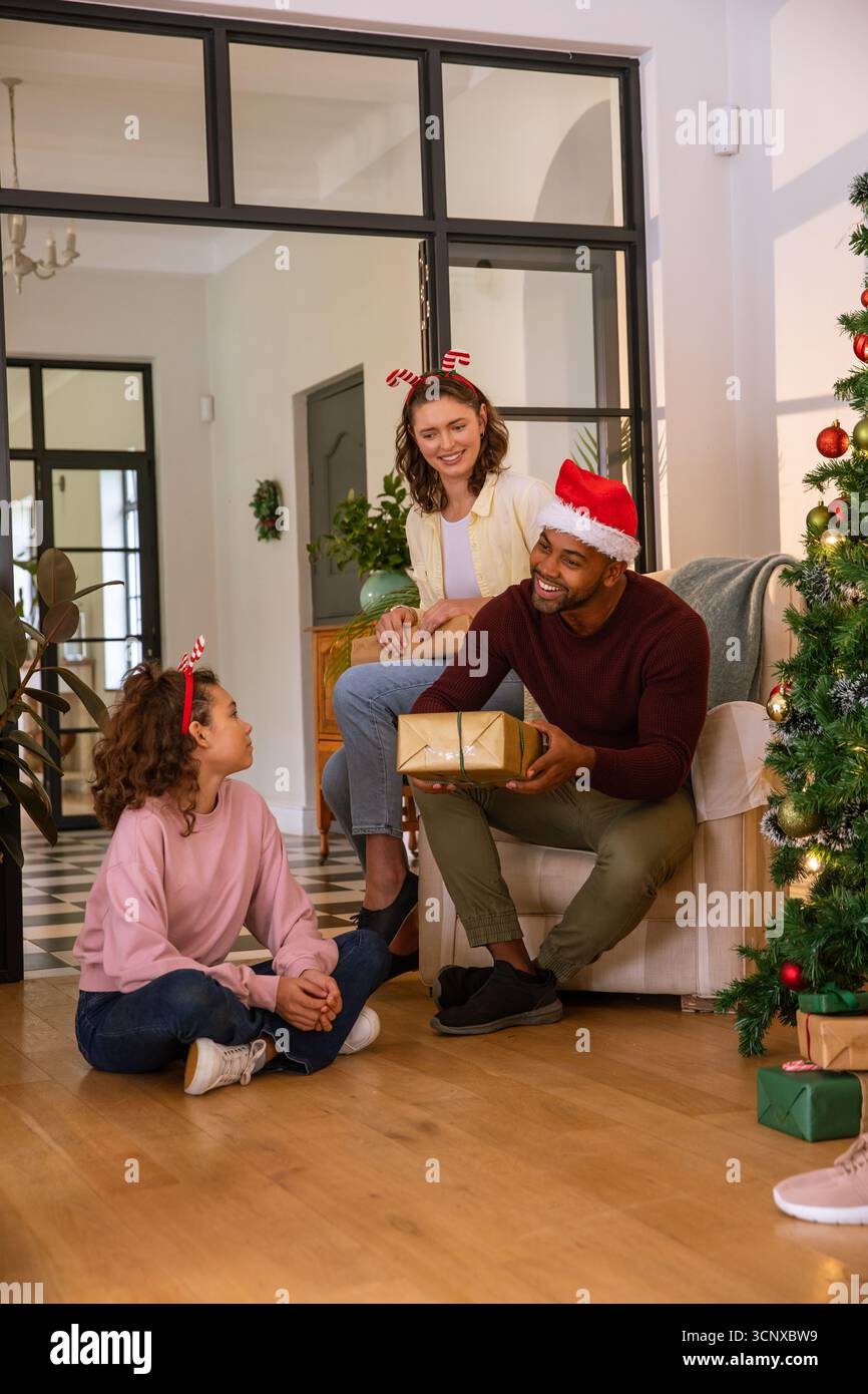 Famille diversifiée échangeant cadeau enveloppé d'or dans des chapeaux de fête dans le salon près de l'arbre de noël Banque D'Images