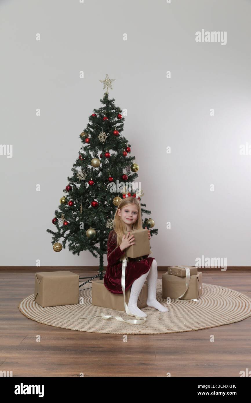 Magie de Noël : jolie fille de 7 ans en robe rouge profitant de la saison des fêtes — emballage de cadeaux de Noël près de l'arbre de fête Banque D'Images