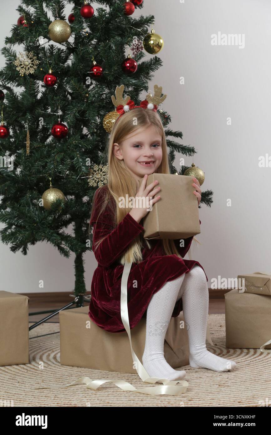 Magie de Noël : jolie fille de 7 ans en robe rouge profitant de la saison des fêtes — emballage de cadeaux de Noël près de l'arbre de fête Banque D'Images