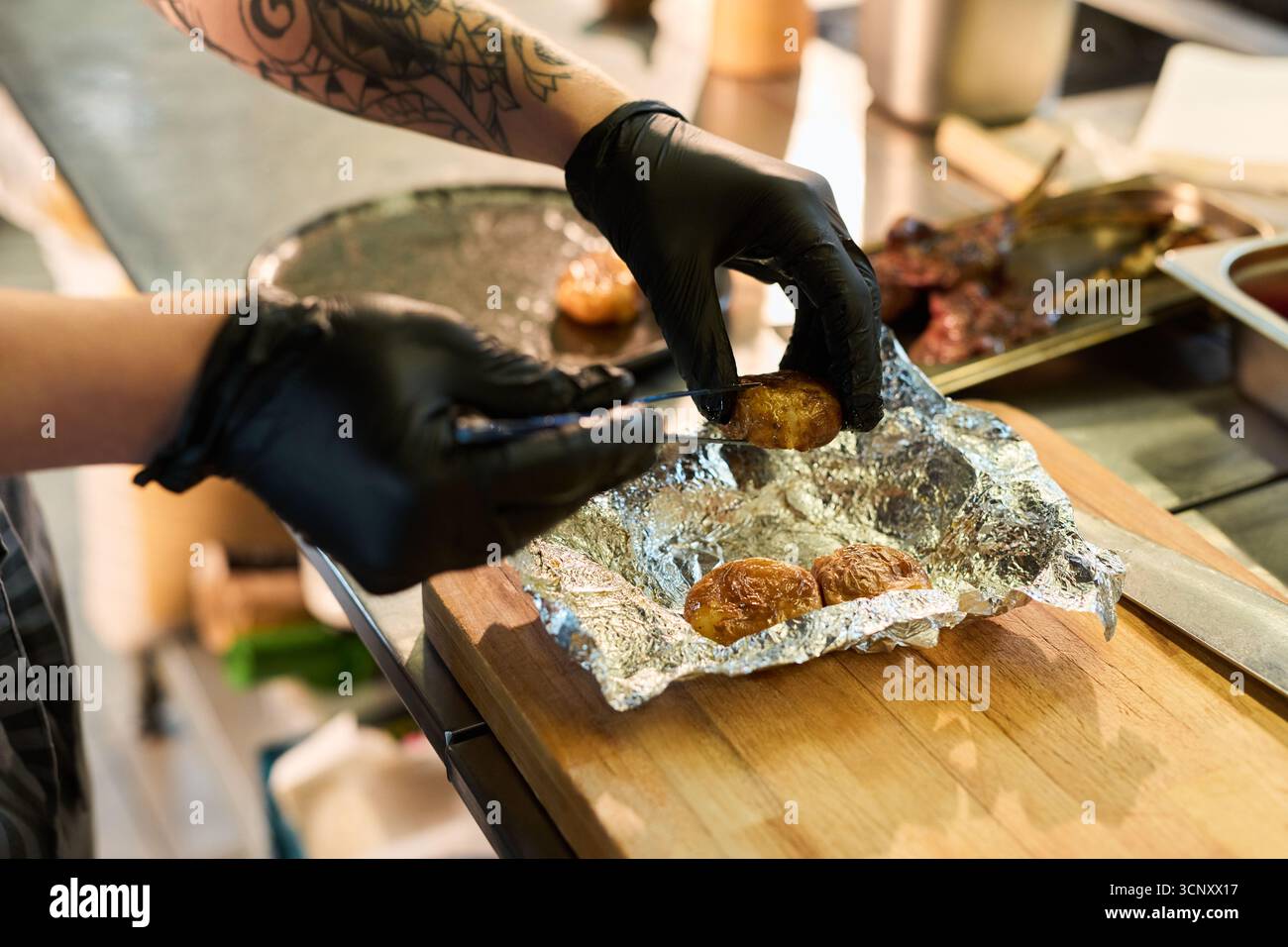 Jeune femme adulte caucasienne au bras tatoué portant des gants noirs préparant des pommes de terre cuites au four en papier d'aluminium sur planche de bois dans la cuisine professionnelle, les mains organisant soigneusement la nourriture pour servir Banque D'Images