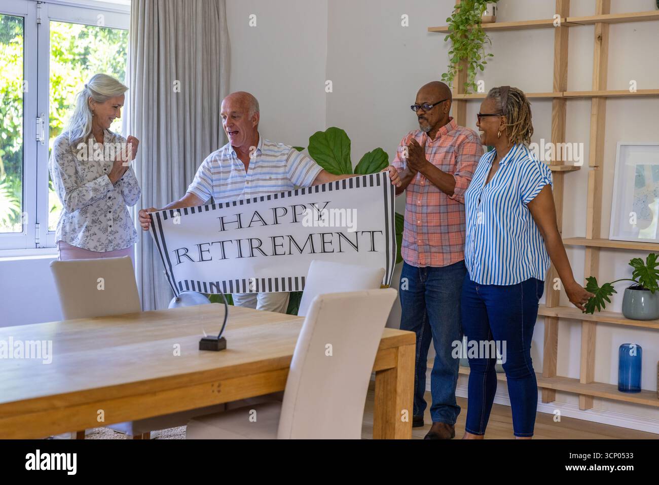 Divers amis seniors tenant la bannière de retraite heureuse, applaudissant dans la salle à manger à la maison avec table Banque D'Images