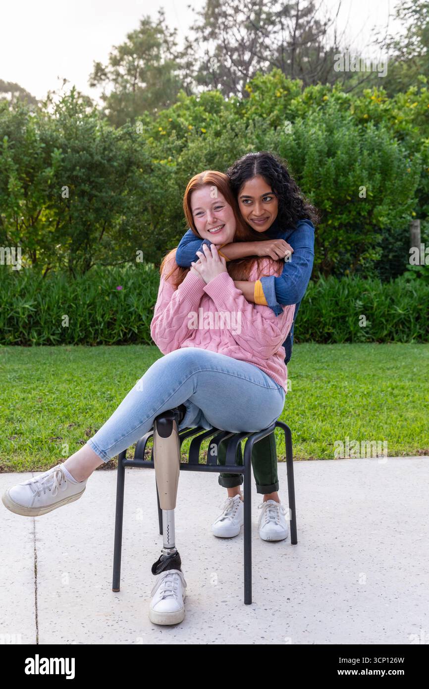 Diverses femmes embrassant sur une chaise en métal sur un patio en béton dans le jardin avec jambe prothétique, baskets Banque D'Images