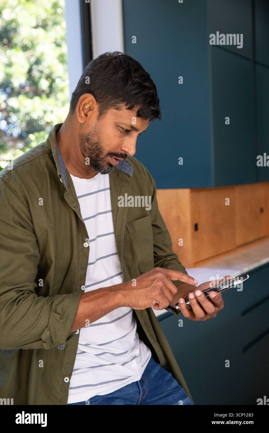 Homme indien appuyé sur le comptoir par la fenêtre, vérifiant smartphone dans la cuisine avec armoires sarcelle Banque D'Images