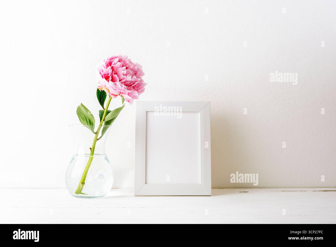 Nature morte minimale avec pivoine rose dans un vase en verre et cadre photo vierge sur une table contre un mur blanc. Maquette. Banque D'Images