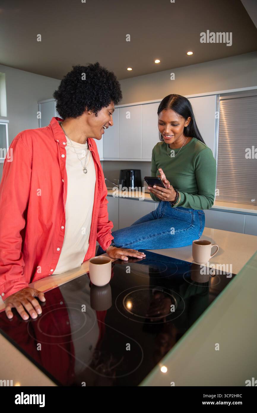 Couple diversifié s'appuyant sur l'îlot de cuisine moderne, partageant le moment smartphone près des tasses, table de cuisson Banque D'Images Couple diversifié s'appuyant sur l'îlot de cuisine moderne, partageant le moment smartphone près des tasses, table de cuisson Banque D'Images