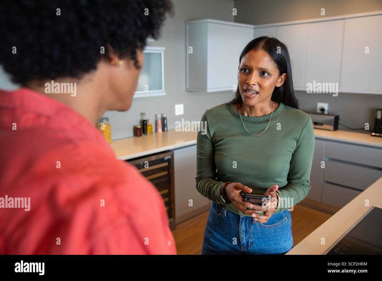 Couple diversifié conversant à l'îlot de cuisine, avec smartphone à côté de pots d'épices et refroidisseur de vin Banque D'Images Couple diversifié conversant à l'îlot de cuisine, avec smartphone à côté de pots d'épices et refroidisseur de vin Banque D'Images