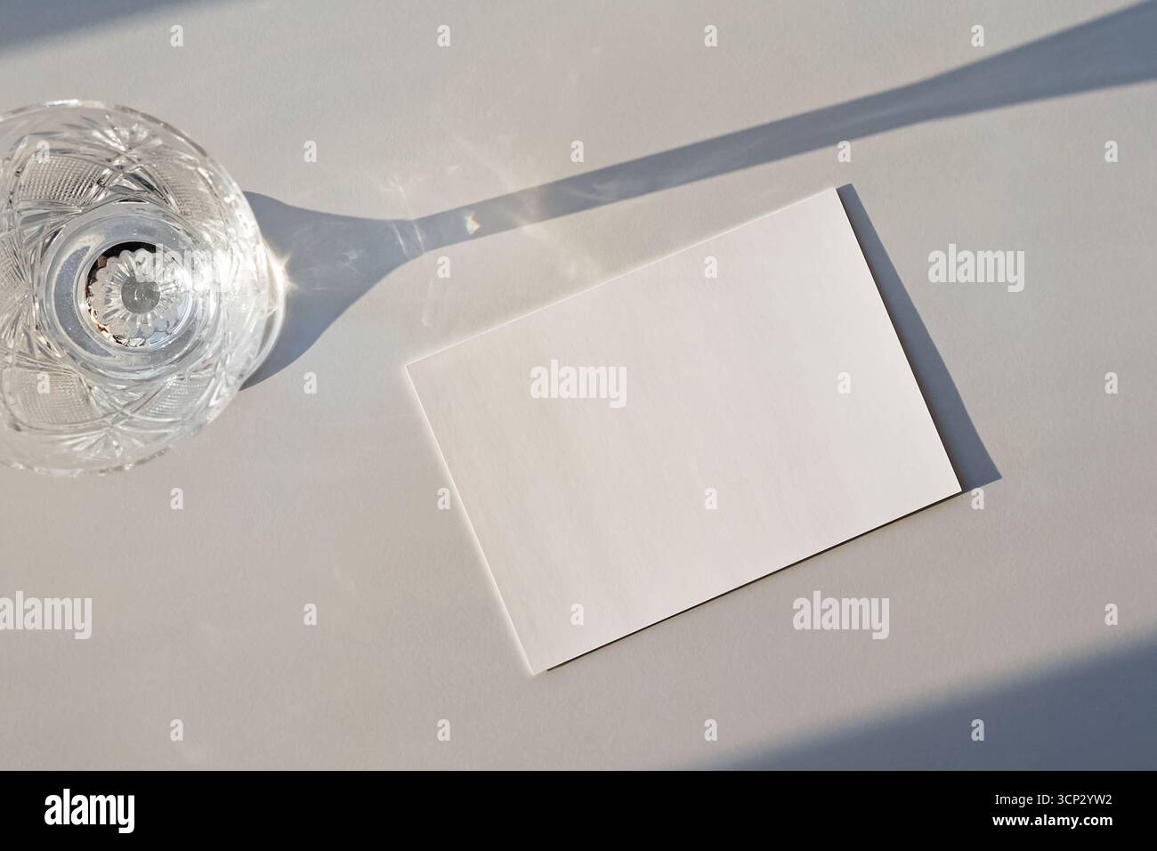 Un verre de cristal projette une longue ombre sur une surface minimaliste, à côté d'une maquette de carte blanche vierge. Banque D'Images