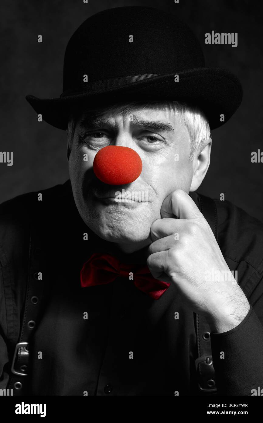 Portrait couleur sélectif dramatique d'un clown triste sur un fond sombre. Un homme âgé avec un nez de clown rouge et un chapeau melon sur la tête. Habillé en blac Banque D'Images