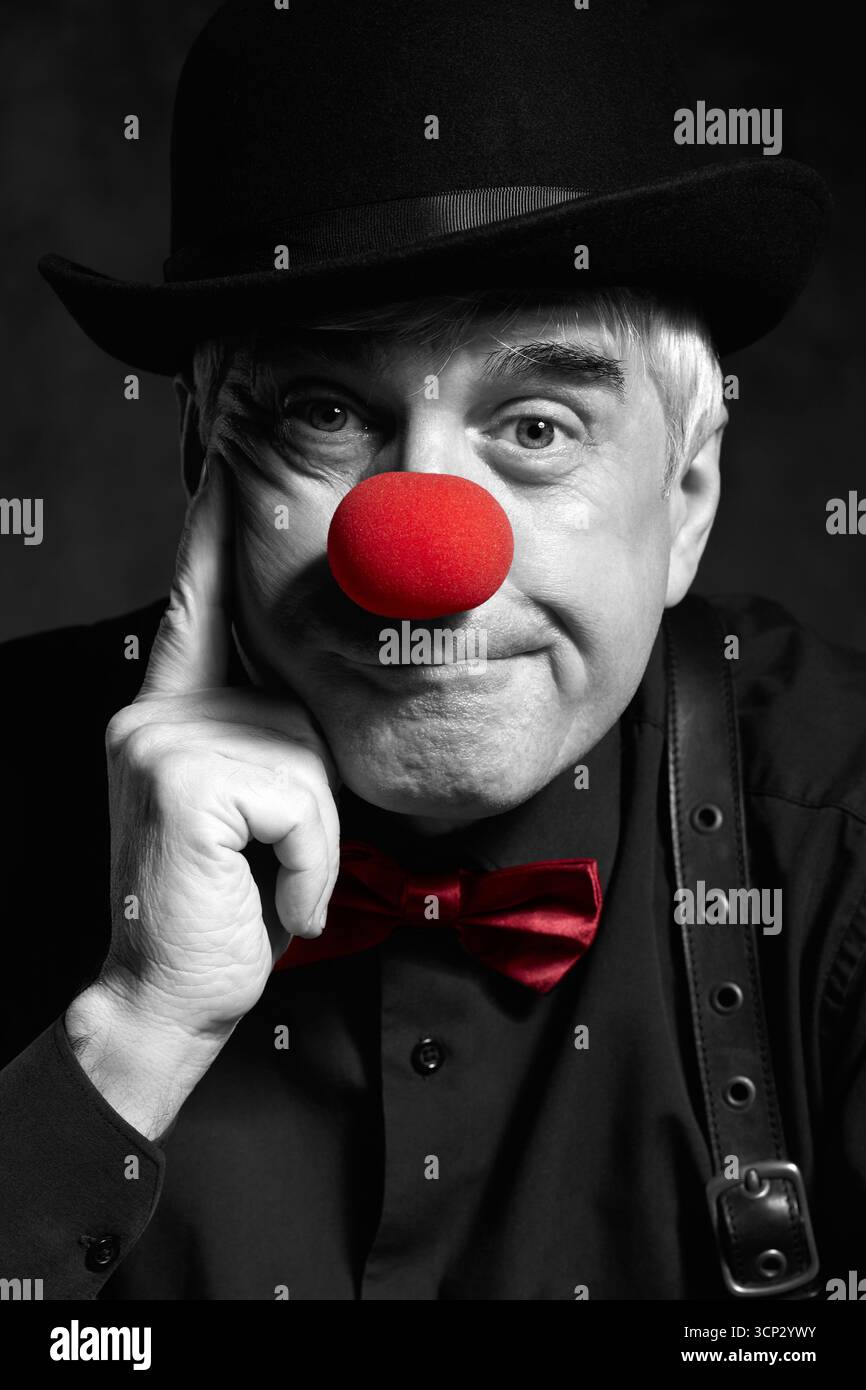 Portrait couleur sélectif dramatique d'un clown triste sur un fond sombre. Un homme âgé avec un nez de clown rouge et un chapeau melon sur la tête. Habillé en blac Banque D'Images