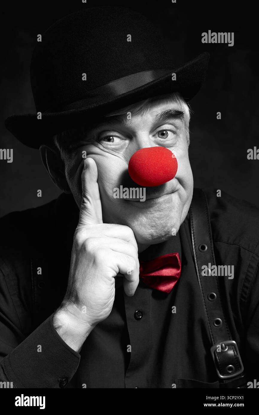 Portrait couleur sélectif dramatique d'un clown triste sur un fond sombre. Un homme âgé avec un nez de clown rouge et un chapeau melon sur la tête. Habillé en blac Banque D'Images