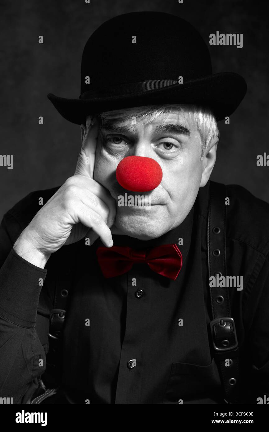 Portrait couleur sélectif dramatique d'un clown triste sur un fond sombre. Un homme âgé avec un nez de clown rouge et un chapeau melon sur la tête. Habillé en blac Banque D'Images