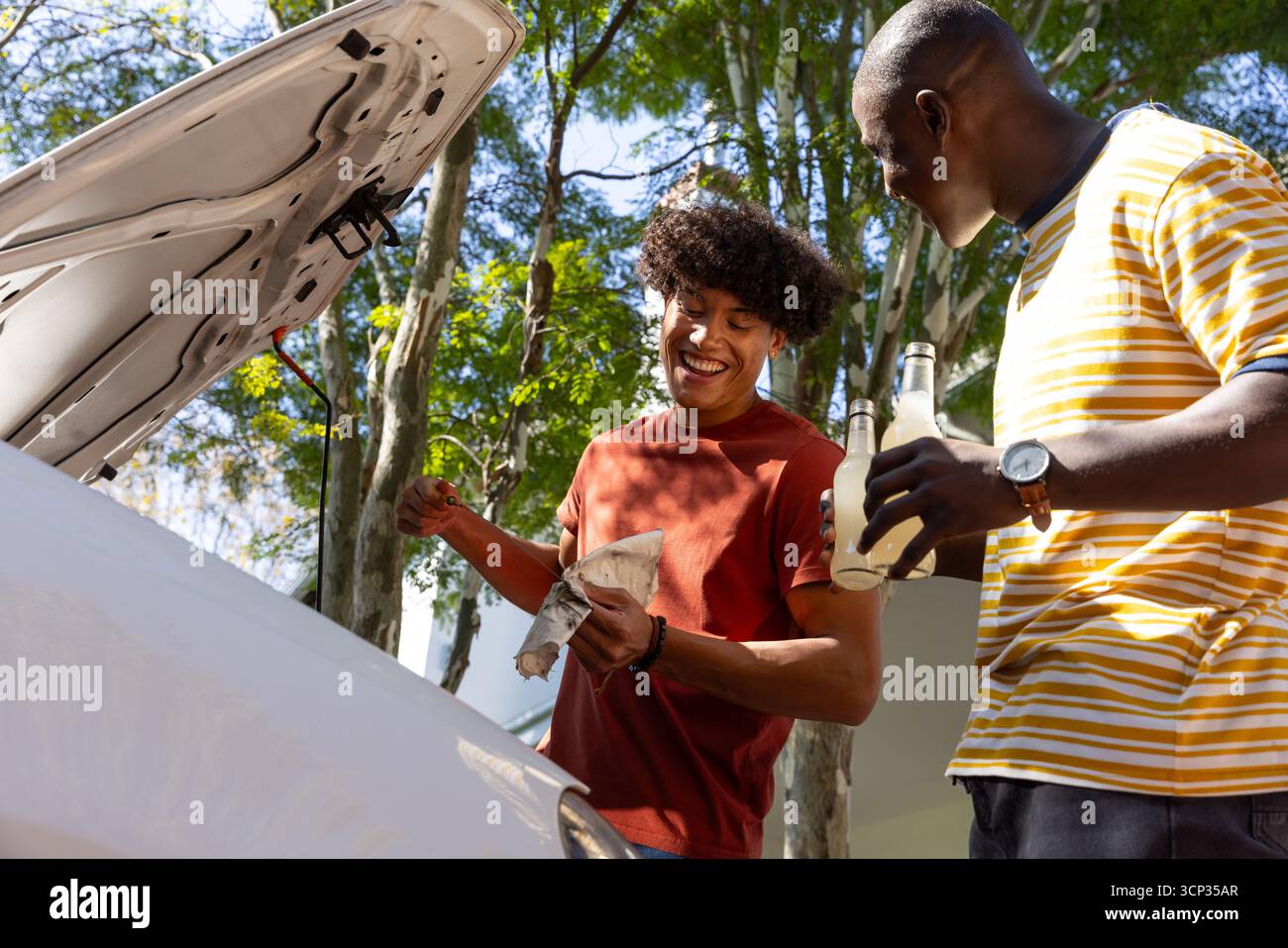 Divers amis masculins inspectant l'huile moteur sous le capot de voiture ouvert dans l'allée, tenant des bières Banque D'Images