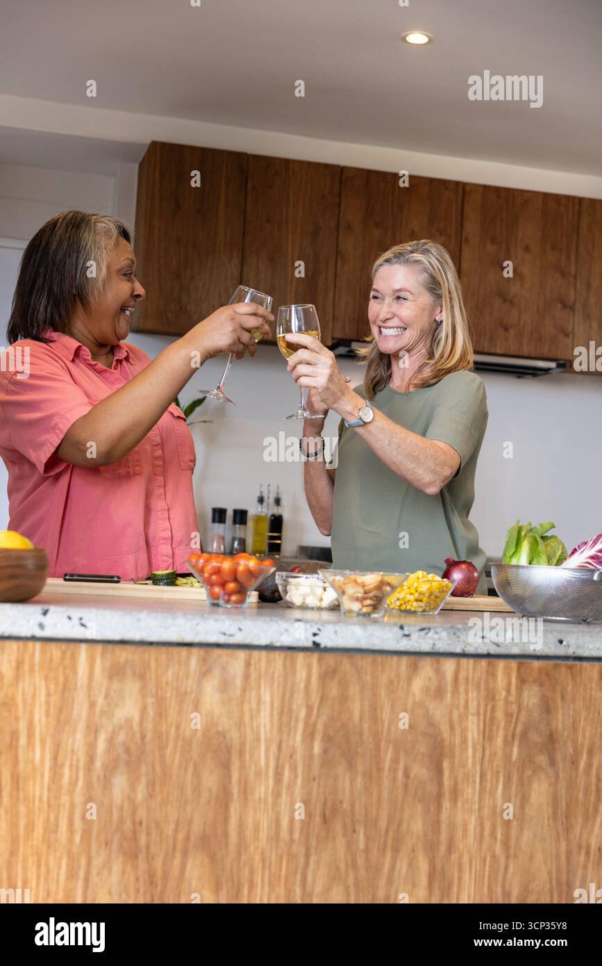 Diverses amies féminines se serrant des verres à vin et mélangeant des ingrédients de salade sur l'îlot de cuisine Banque D'Images