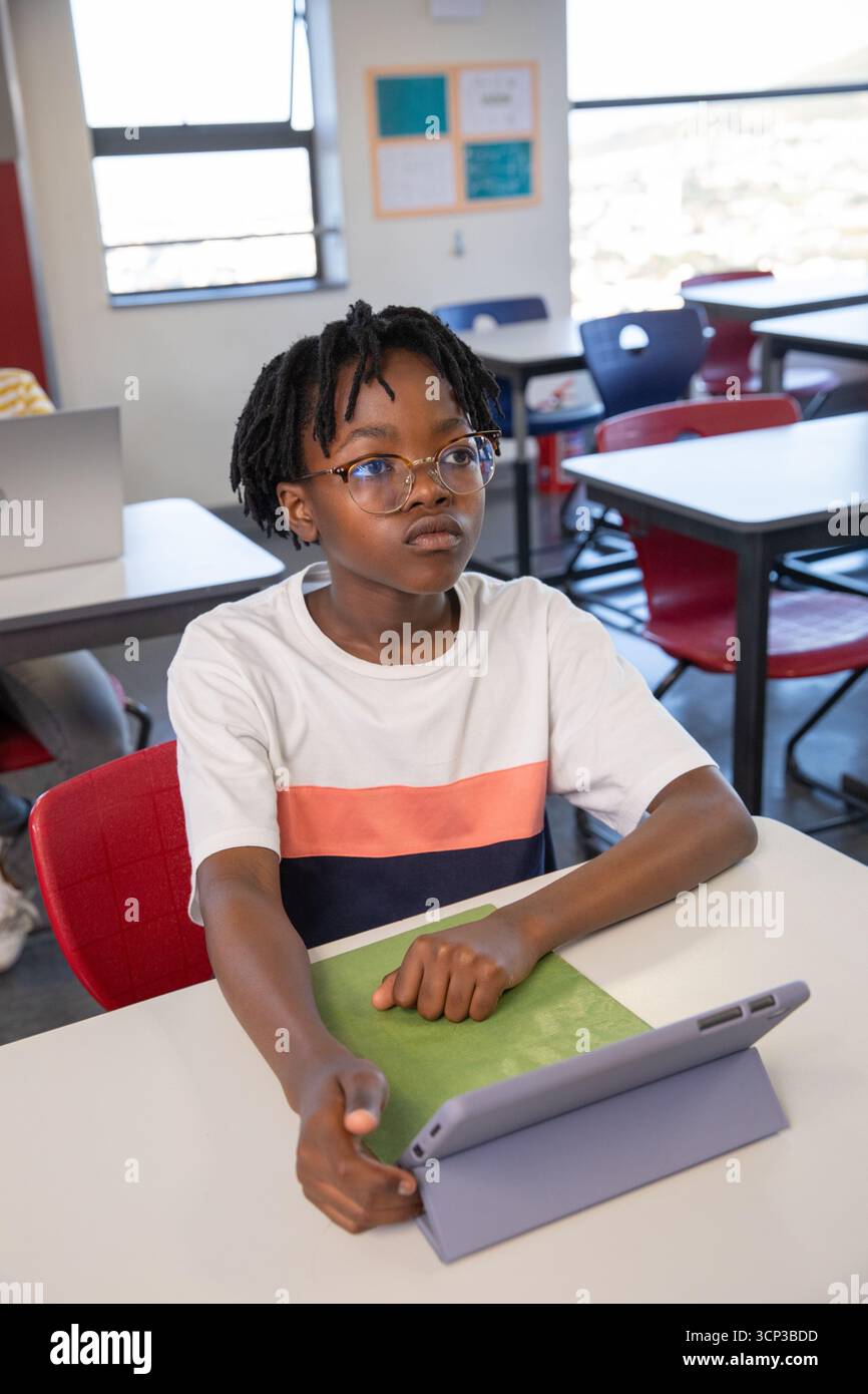 Enfant afro-américain assis dans une chaise rouge au bureau dans la salle de classe en utilisant la tablette dans le cas Banque D'Images