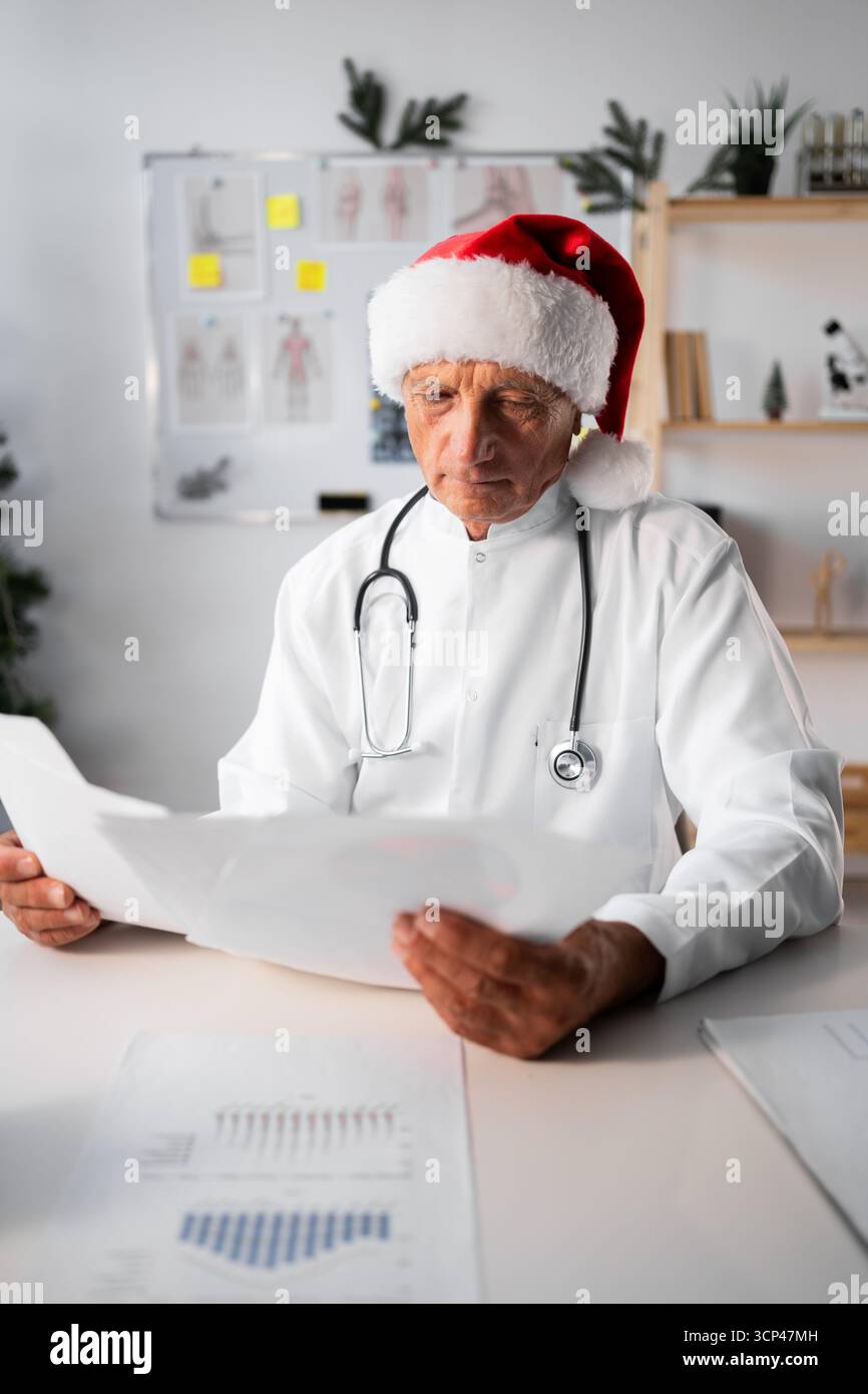 Médecin masculin âgé à Santa Hat avec stéthoscope travaillant avec des documents en clinique. copier l'espace Banque D'Images