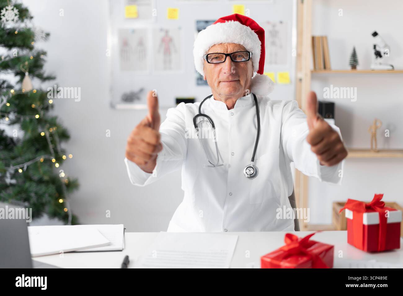 Médecin à la retraite à Santa Hat pointant les pouces vers le haut comme un geste d'accord tout en travaillant dans la clinique. Noël, santé, profession et médecine con Banque D'Images