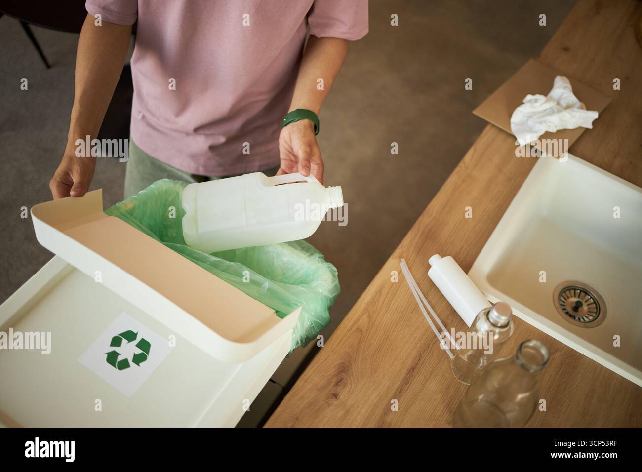 Jeune femme adulte caucasienne jetant une bouteille en plastique dans un bac de recyclage près de l'évier de cuisine, debout au comptoir avec des bouteilles en verre et des déchets de papier visibles sur la surface Banque D'Images