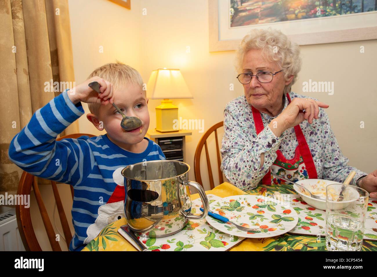 Garçon de 5 ans mange directement dans le pot de crème anglaise tout en étant assis à la table du dîner à côté de sa grand-mère pendant les festivités de Noël, Angleterre Royaume-Uni Banque D'Images