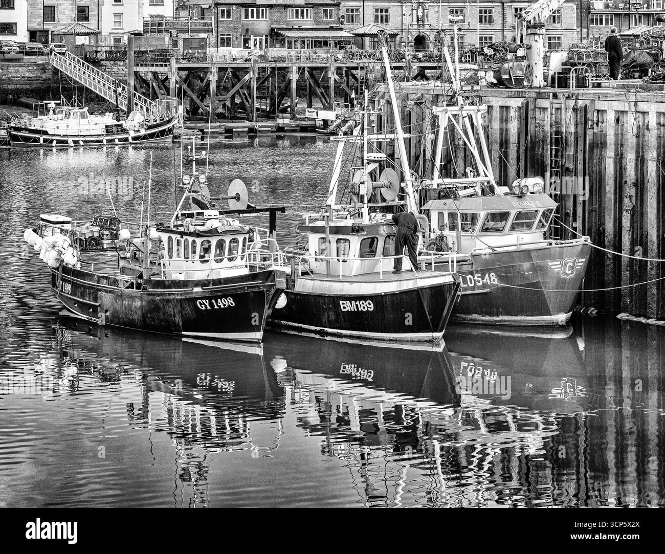 Trois petits bateaux de pêche sont amarrés le long d'un quai et se reflètent dans l'eau. Un canot de sauvetage et des bâtiments sont en arrière-plan. Banque D'Images