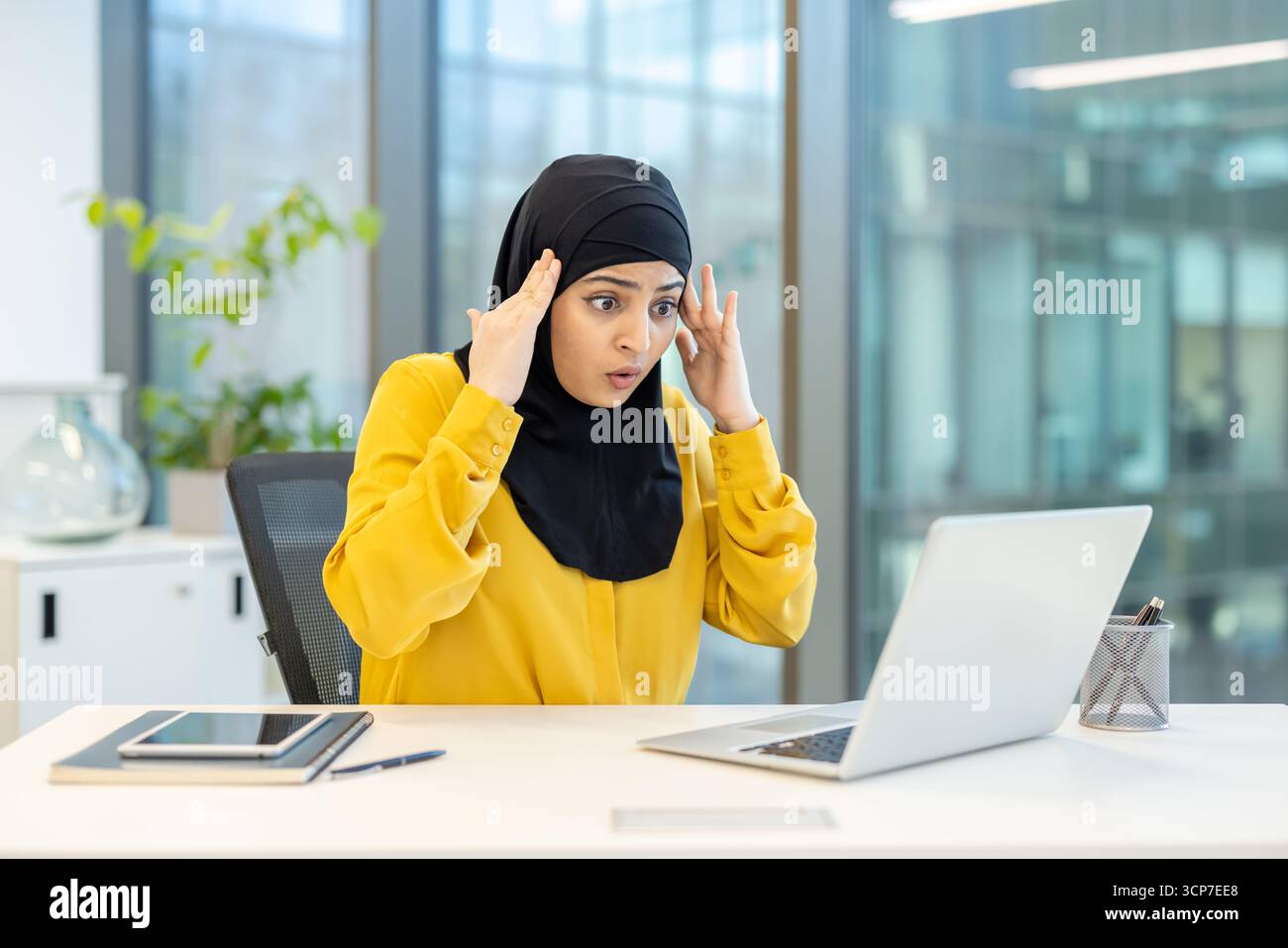 Femme d'affaires musulmane en hijab se sentant choquée et surprise tout en regardant de mauvaises nouvelles sur l'écran de son ordinateur portable, aux prises avec un problème ou une situation inattendue dans un bureau moderne Banque D'Images