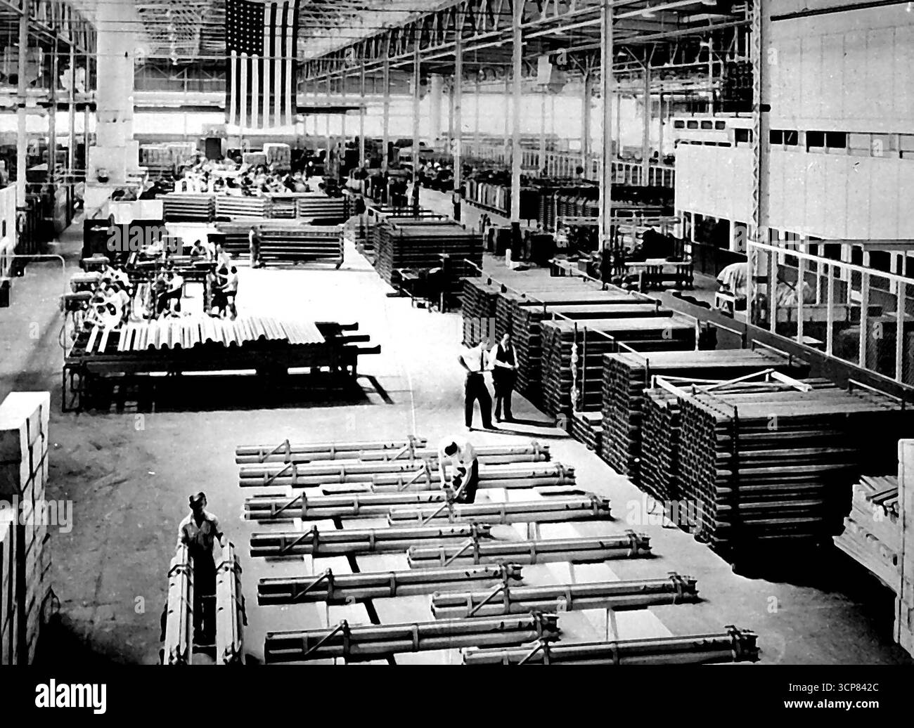 Production de masse de canons à roquettes américains -- des lance-roquettes achevés, prêts à être expédiés sur les fronts de combat, sont placés sur des rayonnages (avant-plan) dans un front de combat, sont placés sur des rayonnages (avant-plan) dans une usine américaine qui produisait autrefois des marchandises en caoutchouc en temps de paix. Les ouvriers à l'arrière de l'atelier utilisent des méthodes de production de masse pour produire des pièces pour les canons, qui sont montés sous les ailes des avions de combat alliés pour une utilisation contre les installations ennemies et les troupes au sol. Le lance-roquettes se compose d'une grappe de trois tubes de tir chacun de trois pieds (3 mètres) de long et 4,5 pouces (114 mm). en diamètre, Banque D'Images