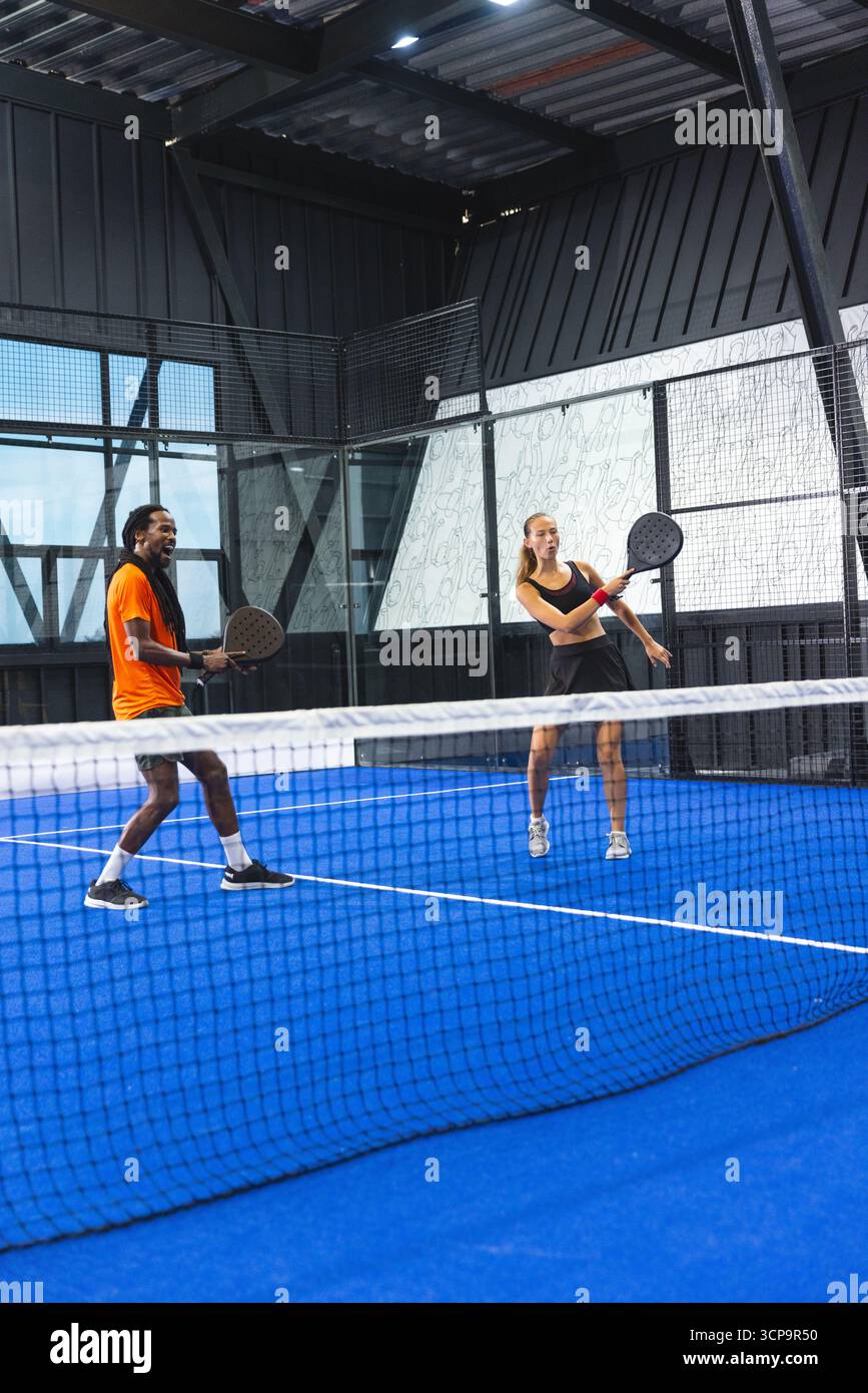 Divers partenaires de padel debout sur la cour bleu vif sous le toit métallique tenant des raquettes par l'escrime Banque D'Images