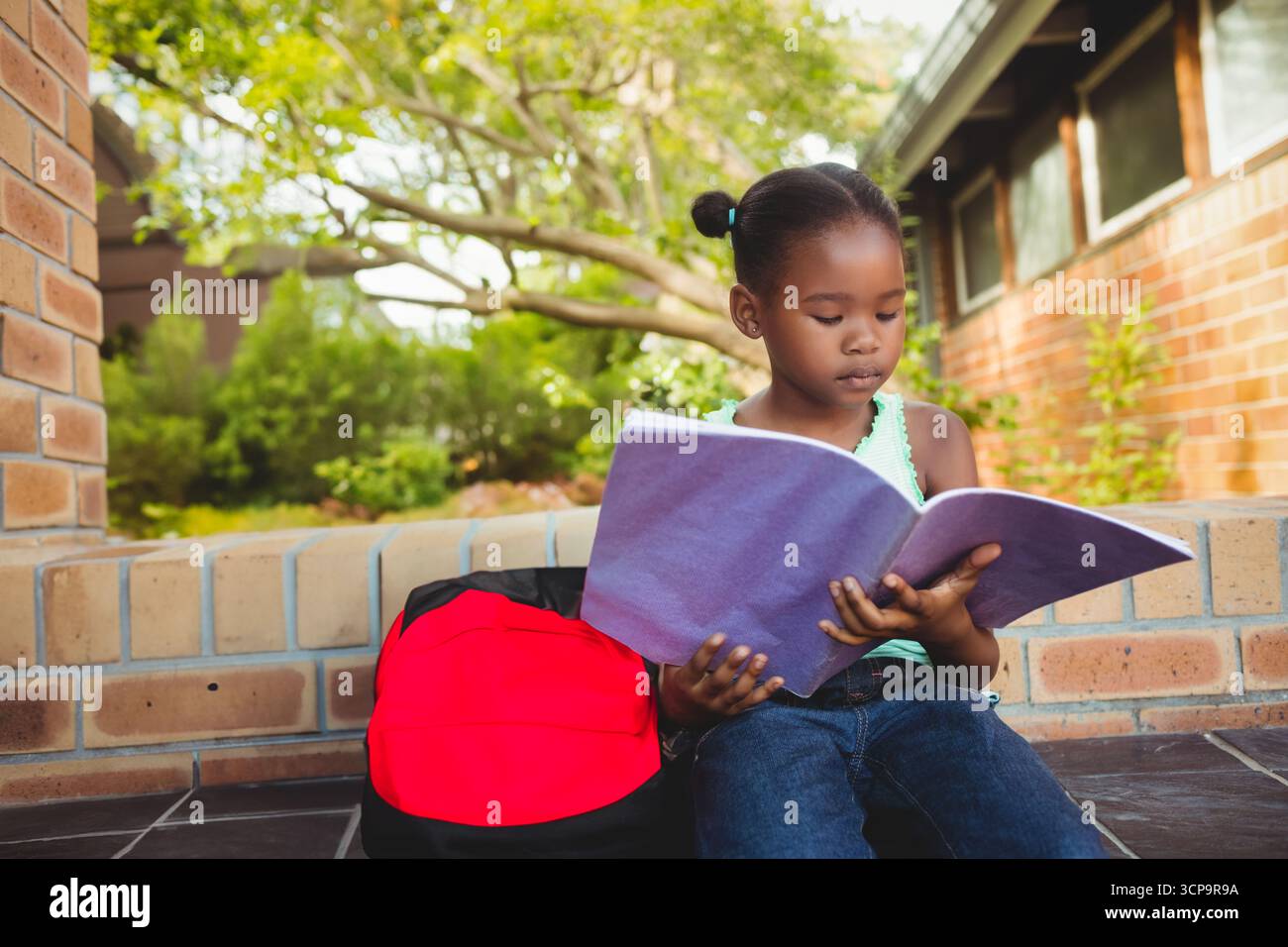 Enfant lisant un grand livre violet sur un rebord bas en briques à l'extérieur du bâtiment, avec sac à dos rouge et noir Banque D'Images