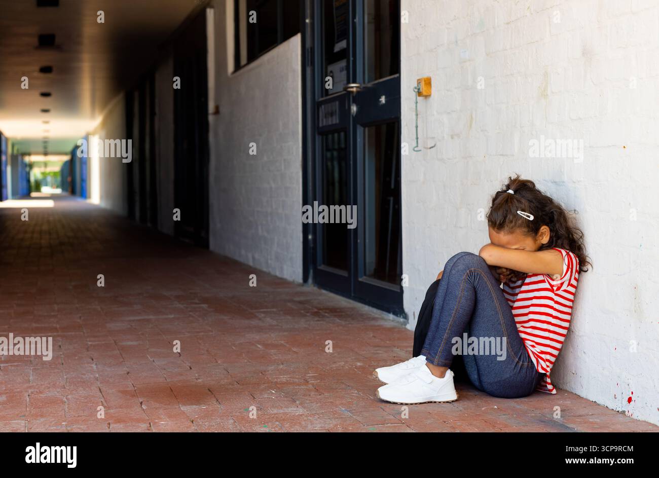 Fille enfant assise sur le sol du couloir de briques rouges dans le couloir couvert portant chemise rayée, espace copie Banque D'Images