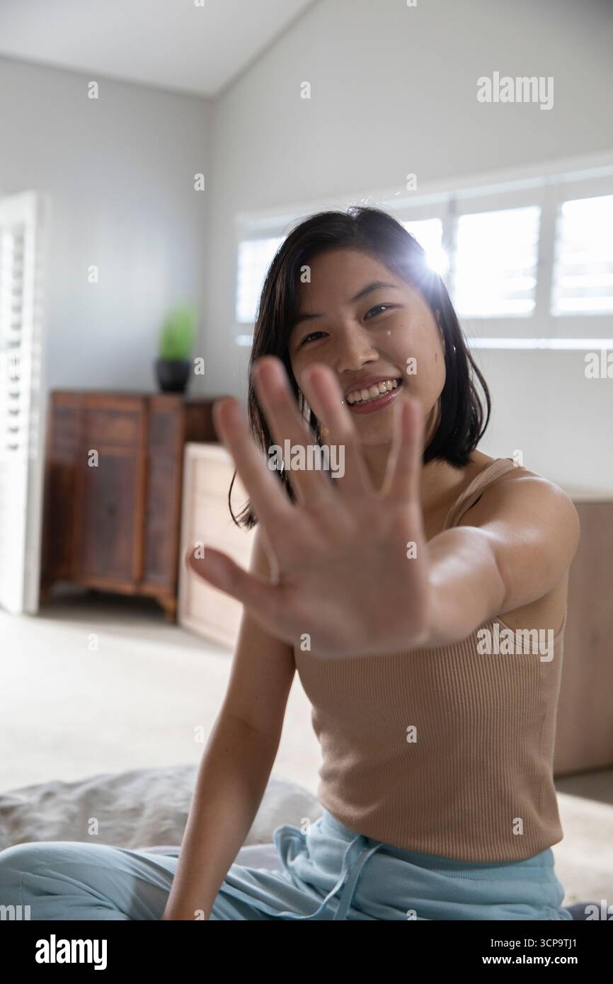 Femme asiatique assise les jambes croisées sur le lit s'étendant la main droite vers la caméra avec la plante en pot Banque D'Images