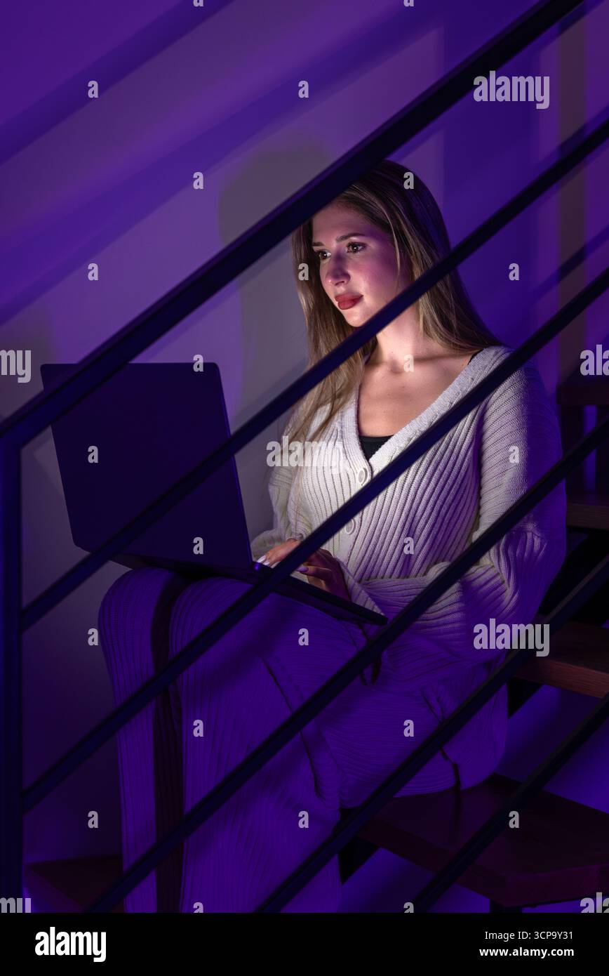 Jeune femme avec ordinateur portable en néon violet sur escalier la nuit. Travail en ligne silencieux. Banque D'Images