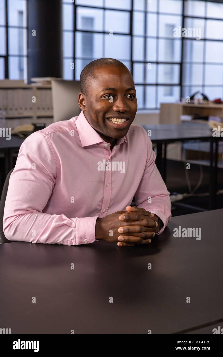 Homme afro-américain en chemise rose souriant et assis au bureau dans le bureau ouvert Banque D'Images