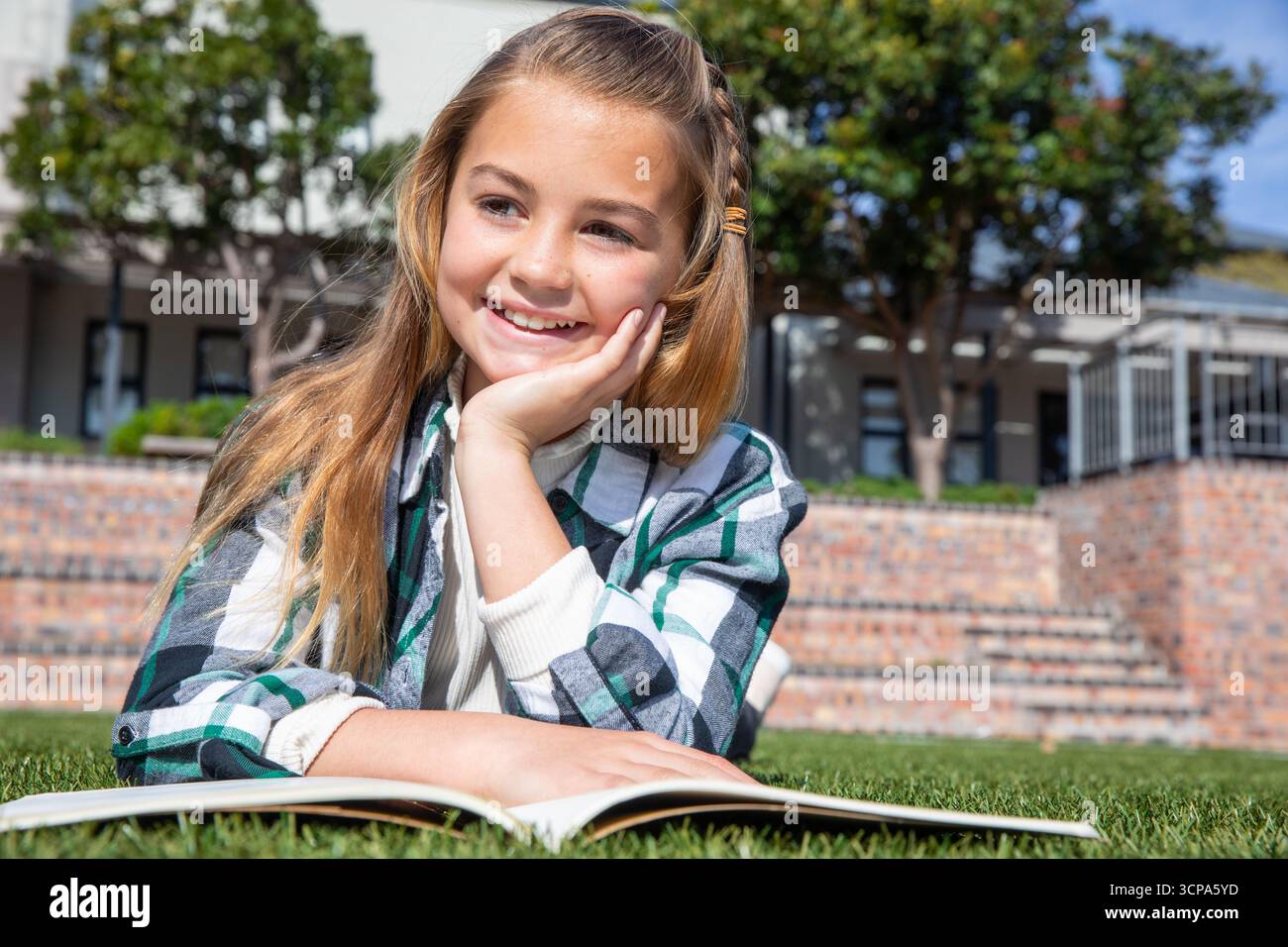 Fille enfant couchée sur l'herbe livre de lecture dans la cour de l'école avec des marches en briques et des arbres Banque D'Images