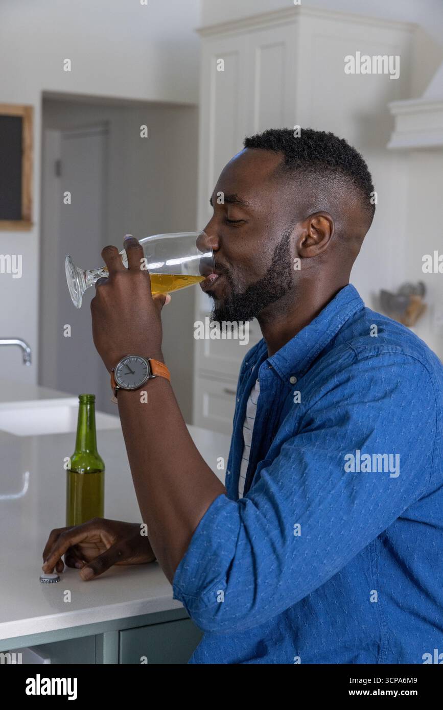 Homme afro-américain buvant boisson à la maison îlot de cuisine avec verre à pied et bouteille Banque D'Images