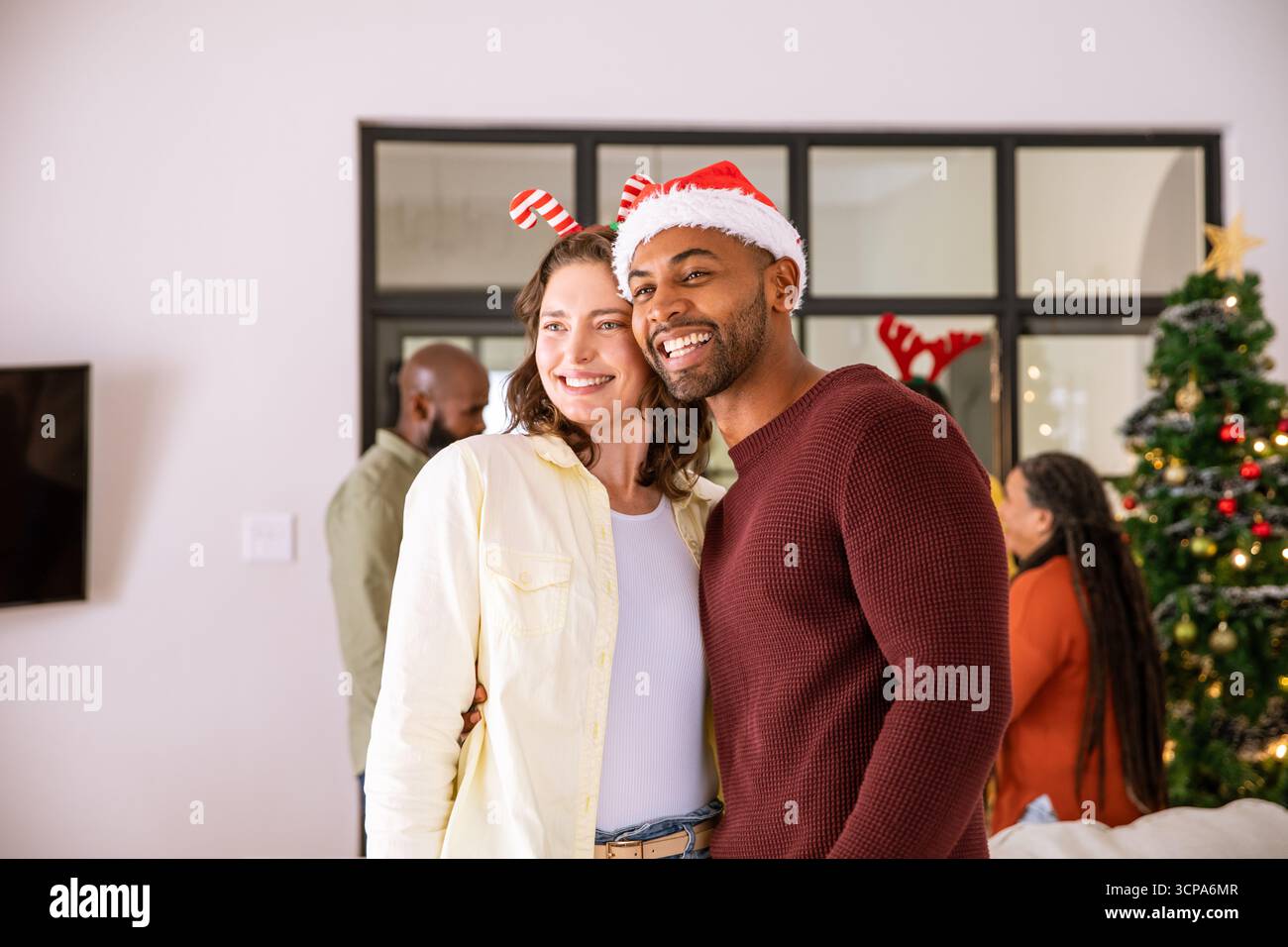 Couple diversifié portant chapeau de père noël, bandeau de canne de bonbons embrassant à la maison près de l'arbre de noël Banque D'Images