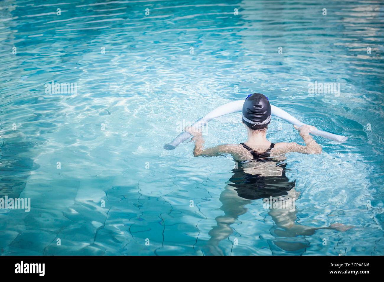 Femme debout à la poitrine dans la piscine extérieure portant maillot de bain et casquette tout en tenant des nouilles en mousse blanche Banque D'Images