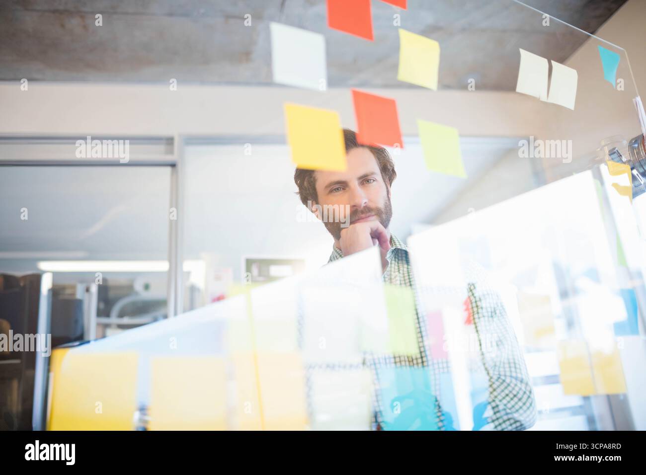 Homme barbu debout derrière un panneau de verre transparent examinant des notes adhésives dans un espace de travail ouvert Banque D'Images
