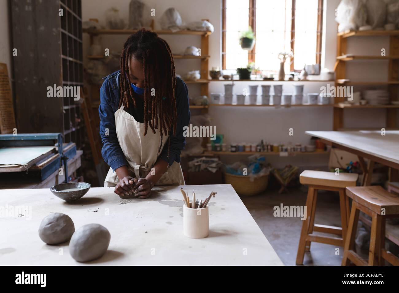 Femme afro-américaine pétrissant l'argile à la table de l'atelier de céramique portant un tablier avec des outils de poterie Banque D'Images Femme afro-américaine pétrissant l'argile à la table de l'atelier de céramique portant un tablier avec des outils de poterie Banque D'Images