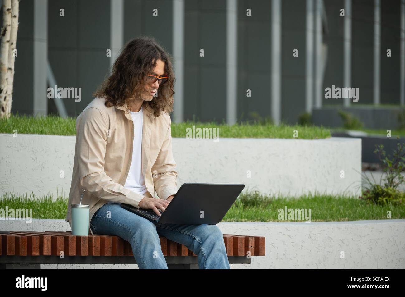 Jeune homme, étudiant, pigiste attrayant est assis sur le banc dans le parc urbain, en utilisant un ordinateur portable pour le travail à distance et l'éducation. À l'extérieur. Banque D'Images