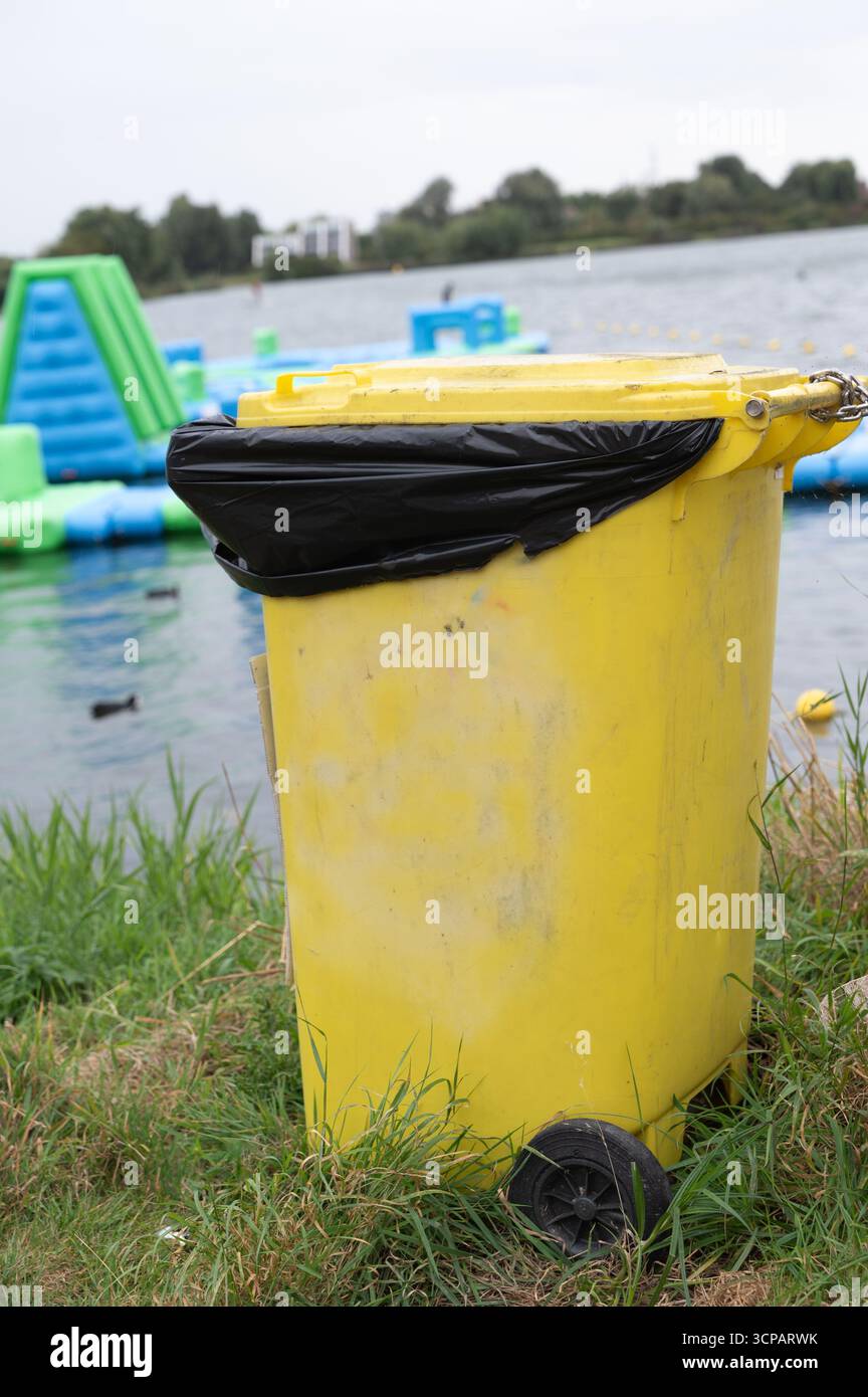 Une poubelle jaune vif est actuellement positionnée dans l'herbe verte luxuriante près d'une belle masse d'eau à proximité, améliorant le paysage Banque D'Images
