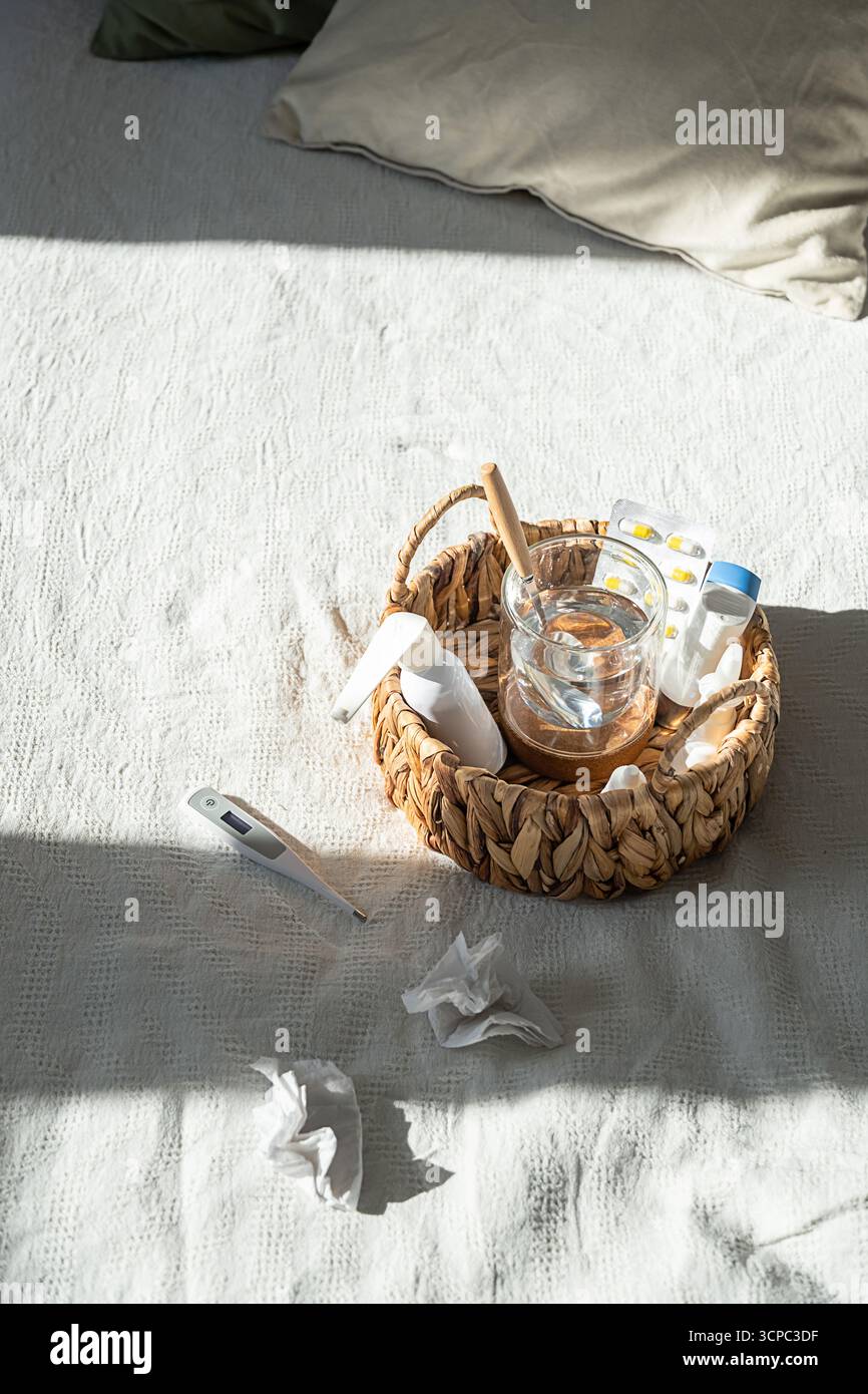 Panier de médicaments avec pilules, verre d'eau, thermomètre et mouchoirs usagés sur lit blanc. Concept de jour de maladie à domicile. Banque D'Images