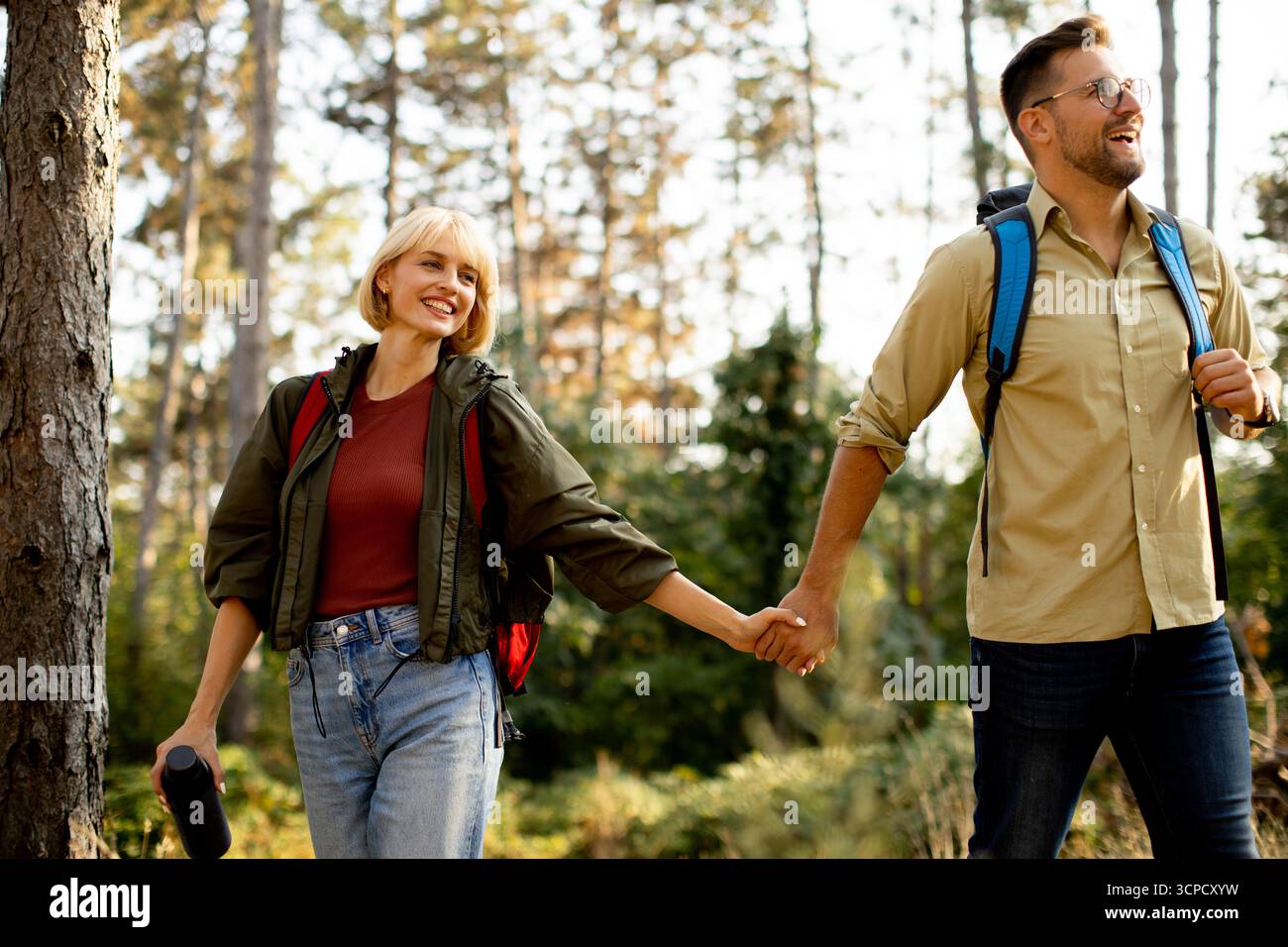 Deux randonneurs explorent une forêt vibrante, souriants et se tenant par la main. Ils sont habillés dans des vêtements de plein air décontractés, profitant d'une journée ensoleillée entourée d'arbres et Banque D'Images
