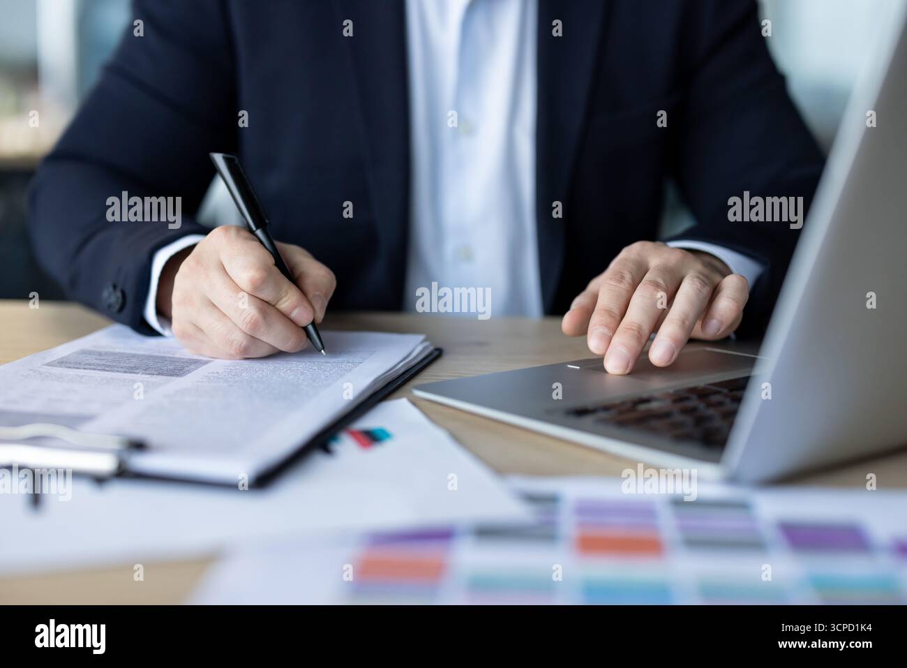 Photo en gros plan des mains d'un jeune homme en costume assis à un bureau dans un bureau, tapant sur un clavier d'ordinateur portable et écrivant un papier. Banque D'Images