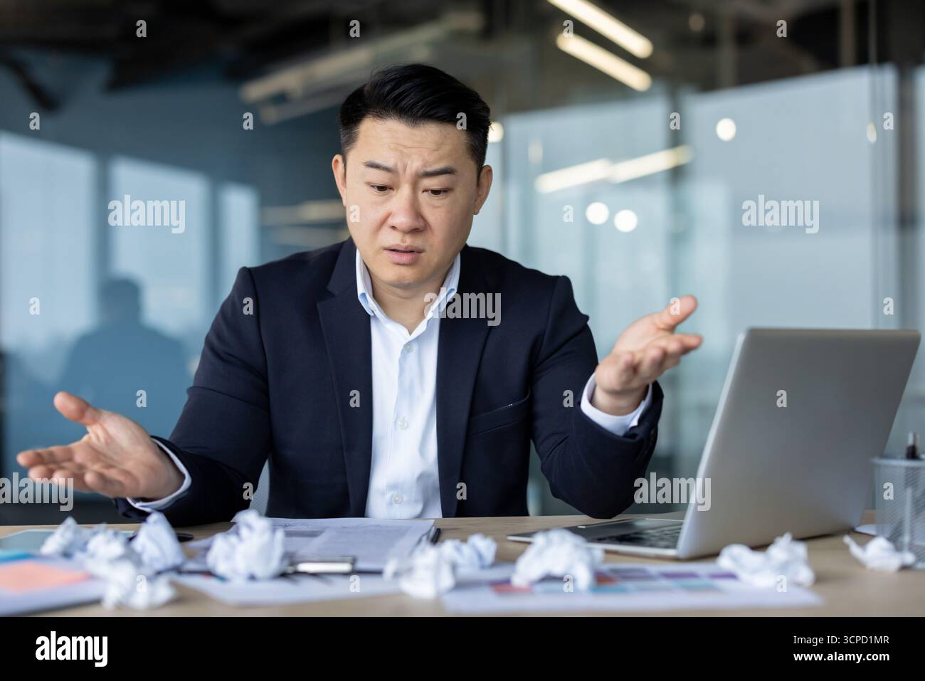 Bouleversé jeune homme asiatique en costume assis au bureau au lieu de travail et regardant déçu en regardant le papier froissé sur la table, écartant les mains. Banque D'Images